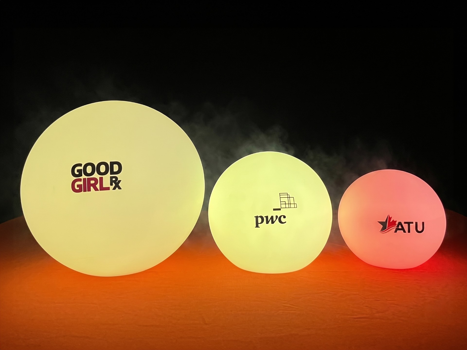 Drei leuchtende Kugeln in verschiedenen Größen mit aufgedruckten Logos von GOOD GIRL, pwc und ATU.